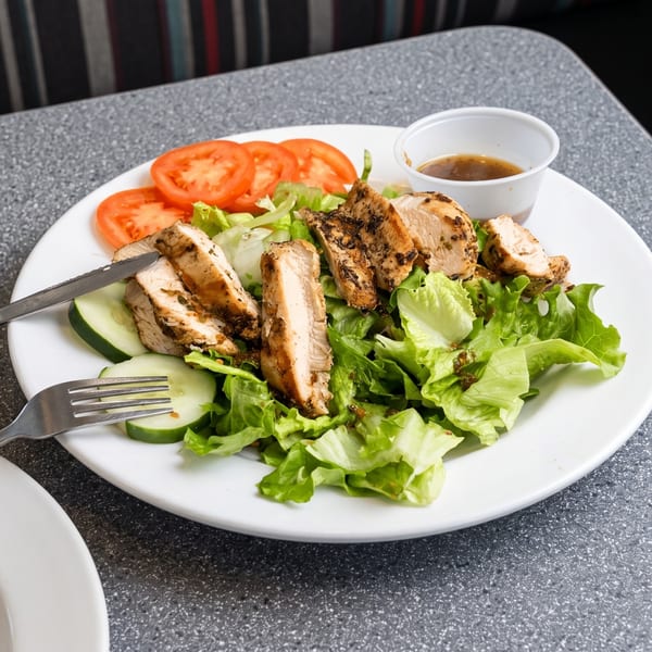 Grilled chicken salad with lettuce, tomatoes, cucumbers, and dressing on a white plate.