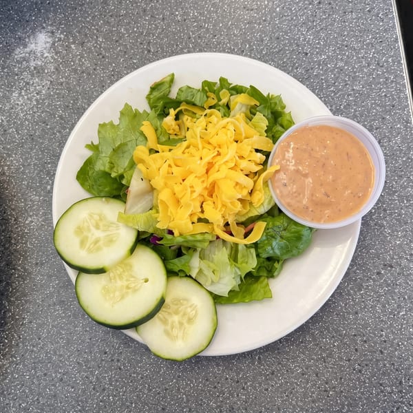 A side salad with lettuce, shredded cheddar cheese, cucumber slices, and a cup of dressing.