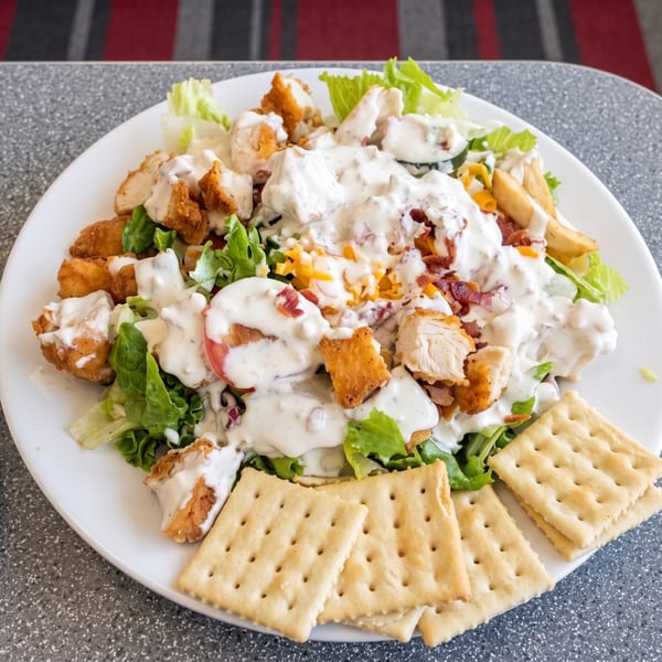 A large salad with fried chicken, bacon, cheese, and ranch dressing, served with crackers.